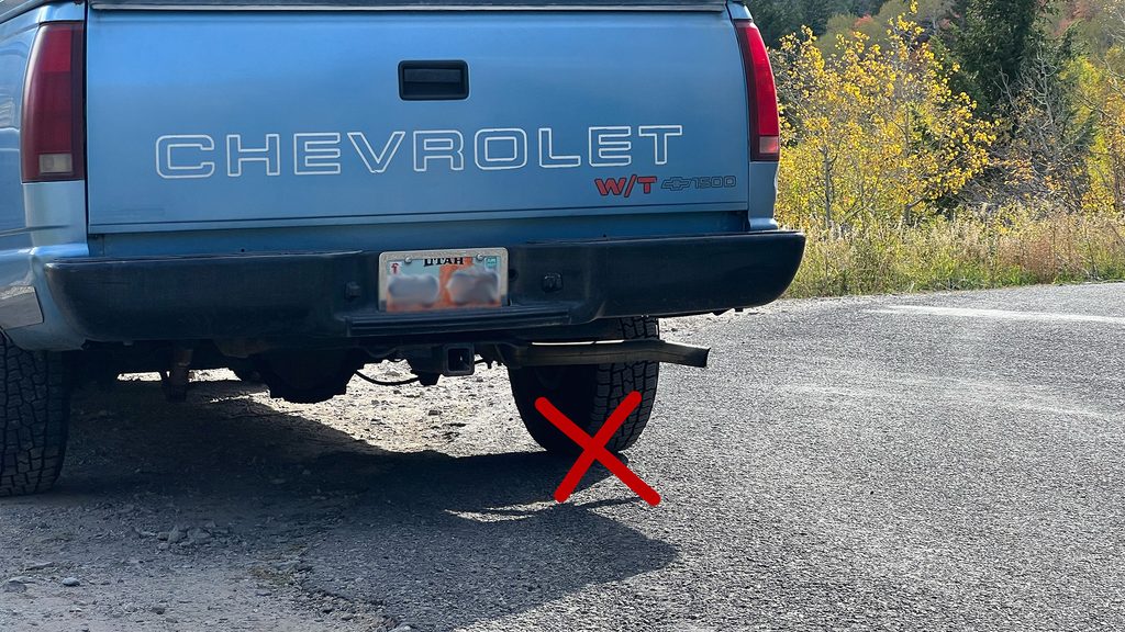 A pickup truck is parked mostly on a dirt roadside pull-off area, but one tire (with a red X drawn on it) is on the pavement of the road.
