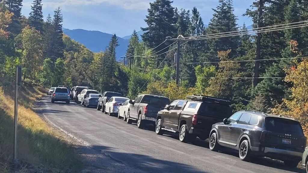 A line of cars is parked on the road, forcing vehicles to cross into oncoming traffic to continue driving. Colorful trees line the road with mountains in the background.