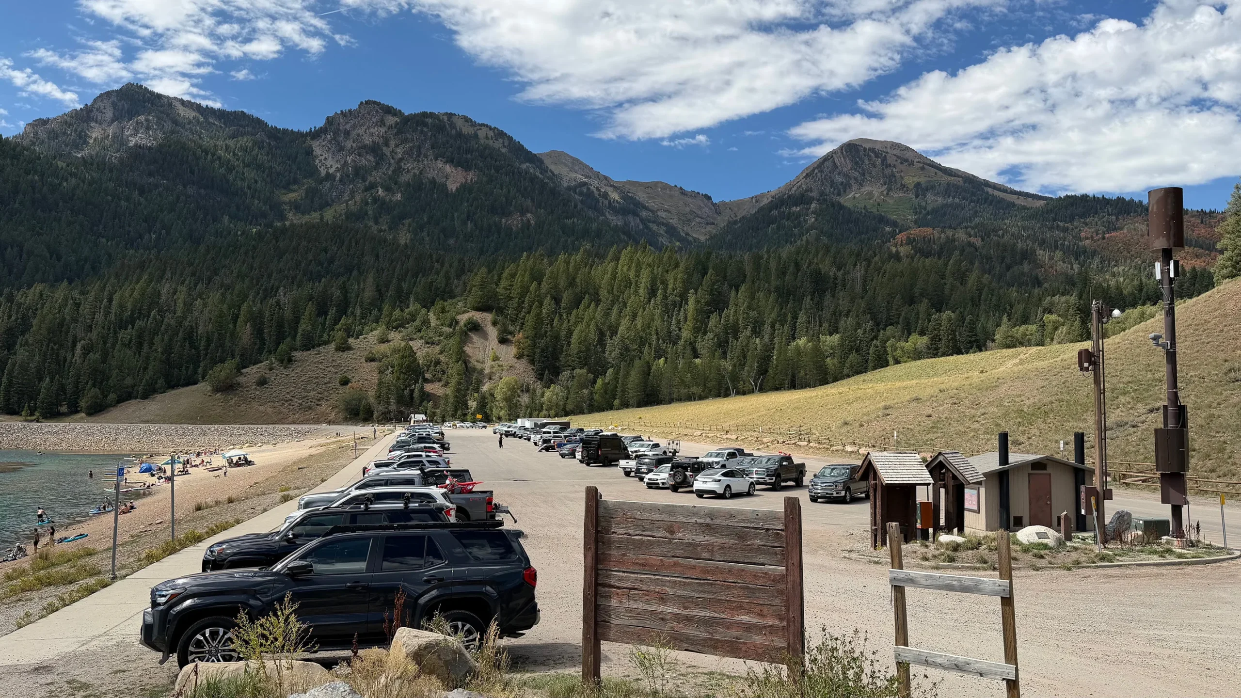 Vehicles fill a large parking lot next to a reservoir in the mountains.